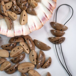 May include: A collection of natural wood half-circle beads with brown and beige tones. The beads are displayed in a seashell and scattered on a white surface.