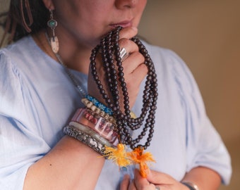 May include: A woman wearing a straw hat and a light blue shirt holds a long strand of dark brown wooden beads. She is wearing several bracelets on her wrist, including a pink and white beaded bracelet, a silver bracelet, and a bracelet with a feather charm.