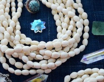 May include: A strand of white oval beads, possibly made of bone or shell, with a turquoise ring and crystal accents. The beads are arranged in a loose pile on a dark blue fabric background.
