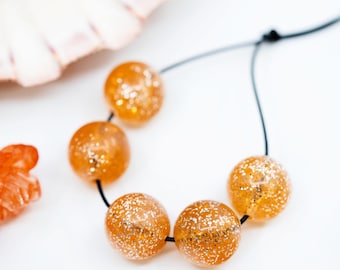 Puede incluir: Un collar hecho con cuentas de vidrio naranja con purpurina dorada. Las cuentas están enhebradas en un cordón negro.