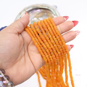 May include: Strands of orange glass beads, each with a cylindrical shape. The beads are held in a hand with red nail polish, and are displayed against a white background. The beads are a vibrant orange color.