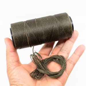 Puede incluir: Una bobina de hilo encerado verde oscuro con un trozo de hilo desenrollado y que descansa sobre una mano.