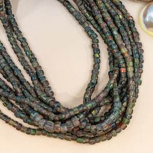 Może przedstawiać: Zbliżenie na wiele sznurów ciemnozielonych i niebieskich szklanych koralików. Koraliki są małe, prostokątne i mają lekko opalizującą jakość. Koraliki ułożone są w kaskadowy wzór, co sugeruje, że są częścią naszyjnika lub podobnej biżuterii.