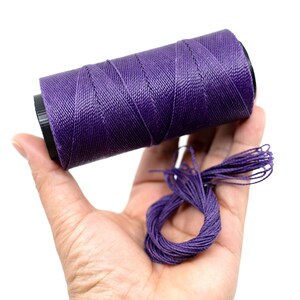 May include: A spool of purple waxed thread for leather crafting. The thread is wound around a black plastic spool and is held in a hand.