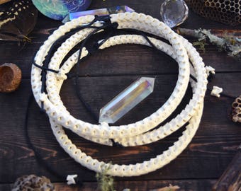 May include: White bone necklace with a black cord, displayed on a wooden surface with a crystal, flowers, and other natural elements.