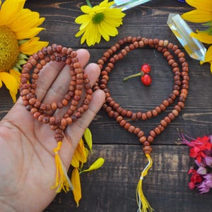 May include: Two brown wooden mala bead necklaces with yellow tassels. The necklaces are on a wooden surface with yellow flowers and other decorative items.