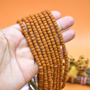 Pode incluir: Um fio de contas de sementes marrons, aproximadamente 10 fios, são mantidos em uma mão. As contas são pequenas e redondas, e o fio é longo.