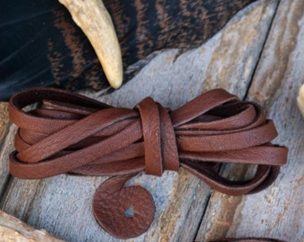 Può includere: Due cordoni di cuoio marrone con un pezzo rotondo di cuoio ad ogni estremità. I cordoni sono su una superficie di legno con due pezzi di corna.