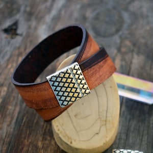 May include: A brown leather bracelet with a silver magnetic clasp. The clasp has a geometric pattern. Two additional silver clasps with different patterns are shown on a wooden surface.