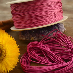 May include: A spool of pink waxed cotton cord, wound around a white spool. The cord is also shown in a loose pile, with the ends of the cord visible.