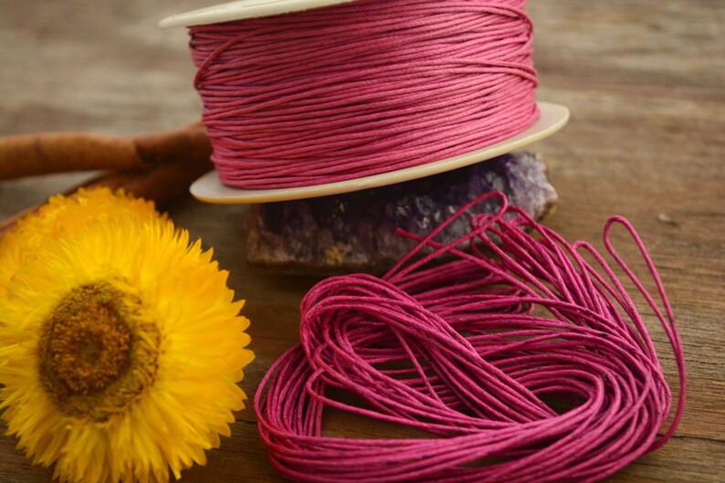May include: A spool of pink waxed cotton cord, wound around a white spool. The cord is also shown in a loose pile, with the ends of the cord visible.