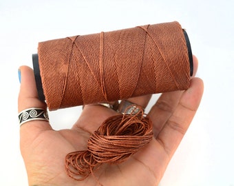 May include: A spool of brown thread, tightly wound, with a small bundle of loose thread in the palm. The thread is a warm, earthy tone, ideal for crafts and sewing projects. The spool is held in a hand against a white background.