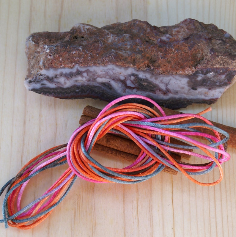 May include: A natural stone with brown and white layers, colorful cords in shades of pink, orange, and gray, and two cinnamon sticks on a light wood surface. The cords are bundled together, creating a vibrant contrast.