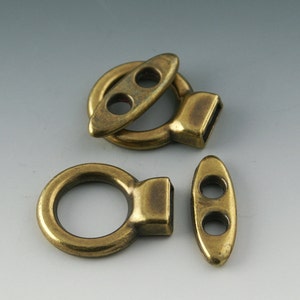 May include: Three antique brass-colored clasps. One is a ring with a rectangular end, another is a ring with an oval piece, and the third is an oval piece with two holes. These clasps are likely for jewelry or bags.