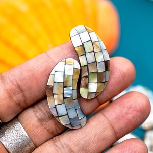 May include: A pair of shell earrings with a mosaic design in shades of white, brown, and beige. The earrings are shaped like a teardrop and have a smooth, polished finish.