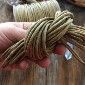 May include: A bundle of olive green cord, tightly coiled and held in a hand. The cord appears to be made of a soft, flexible material. A spool of the same cord is visible in the background, along with a crystal.