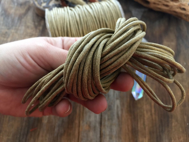 May include: A bundle of olive green cord, tightly coiled and held in a hand. The cord appears to be made of a soft, flexible material. A spool of the same cord is visible in the background, along with a crystal.