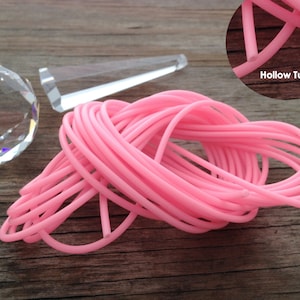 Peut inclure: Gros plan sur un tube creux rose, montrant l'intérieur du tube. Le tube est enroulé et disposé en pile sur une surface en bois. Le texte "Hollow Tubing" est visible sur l'image.