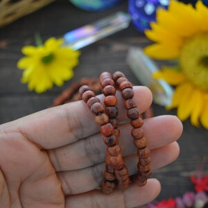 May include: A close-up of a hand holding a brown wooden mala bead necklace. The necklace is made of small, round beads and is strung on a thin cord. The beads are arranged in a circular pattern, and the necklace is worn around the wrist.