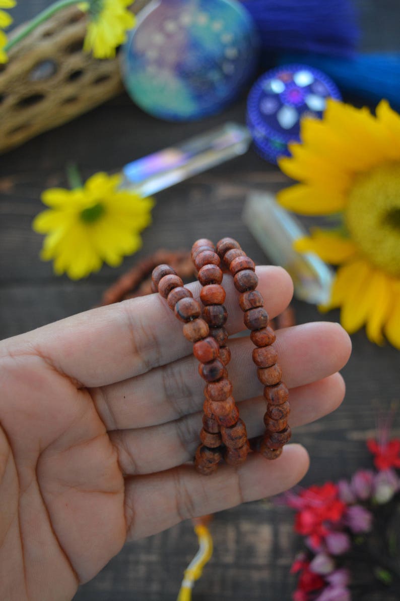 May include: A close-up of a hand holding a brown wooden mala bead necklace. The necklace is made of small, round beads and is strung on a thin cord. The beads are arranged in a circular pattern, and the necklace is worn around the wrist.