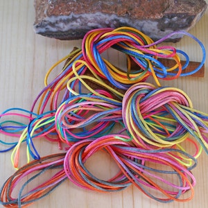 May include: A collection of colorful, tightly wound cords in various hues, including red, blue, yellow, and pink. The cords are arranged on a light wooden surface, with a piece of stone and a cinnamon stick in the background. The cords appear to be made of a smooth, shiny material.