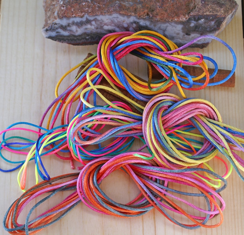 May include: A collection of colorful, tightly wound cords in various hues, including red, blue, yellow, and pink. The cords are arranged on a light wooden surface, with a piece of stone and a cinnamon stick in the background. The cords appear to be made of a smooth, shiny material.