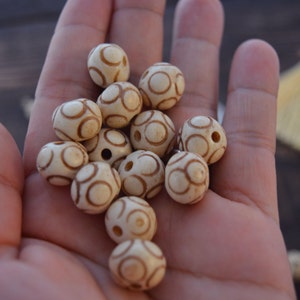 May include: A collection of white wooden beads with brown circular patterns. The beads are arranged in a pile in a person's hand.