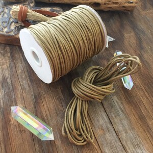 May include: A spool of tan-colored cord sits next to a bundle of the same cord, tied in a knot. Two iridescent crystal points are also present. The cord is likely used for crafting or jewelry making. The items are on a rustic wooden surface.