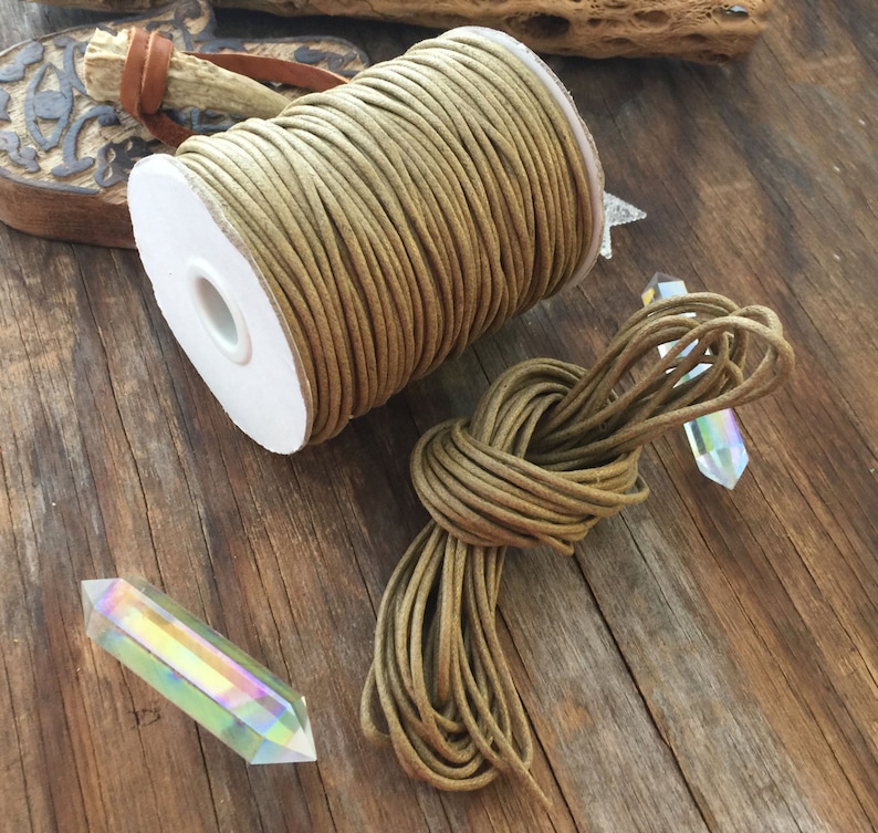 May include: A spool of tan-colored cord sits next to a bundle of the same cord, tied in a knot. Two iridescent crystal points are also present. The cord is likely used for crafting or jewelry making. The items are on a rustic wooden surface.