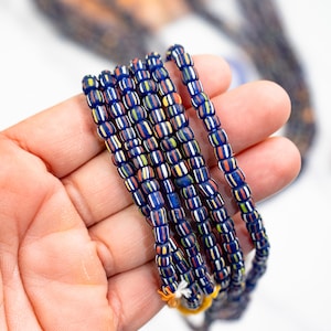 May include: A close-up of a hand holding a stack of blue, white, red, and yellow striped glass beads.