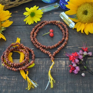May include: Two wooden mala bead necklaces with yellow tassels on a dark wood surface. The necklaces are surrounded by sunflowers, a small bouquet of pink and red flowers, and crystals.