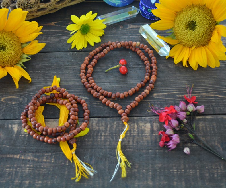 May include: Two wooden mala bead necklaces with yellow tassels on a dark wood surface. The necklaces are surrounded by sunflowers, a small bouquet of pink and red flowers, and crystals.