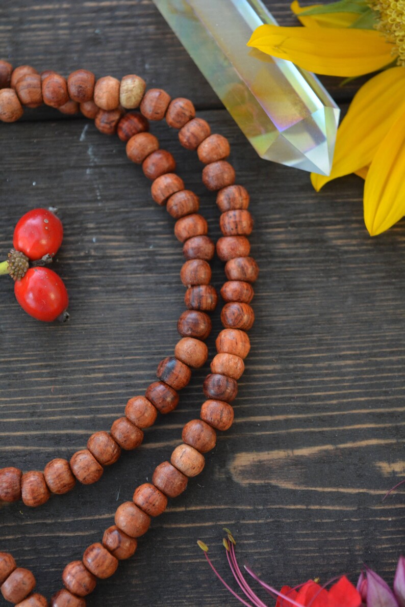 May include: A close-up of a wooden mala necklace with brown beads. The necklace is lying on a dark wooden surface.