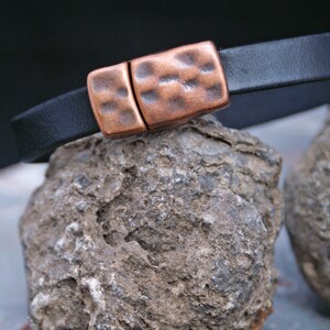 Puede incluir: Un brazalete de cuero negro con un cierre de cobre martillado. El brazalete est&aacute; apoyado en una roca gris y rugosa.