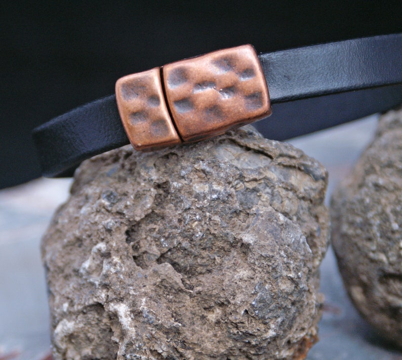 Puede incluir: Un brazalete de cuero negro con un cierre de cobre martillado. El brazalete est&aacute; apoyado en una roca gris y rugosa.