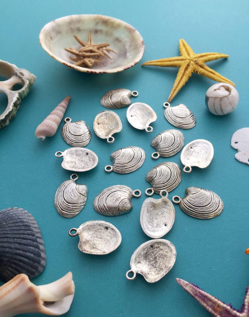 May include: A collection of silver-toned metal clam shell charms with a textured surface. The charms have a loop at the top for attaching to jewelry or other crafts.