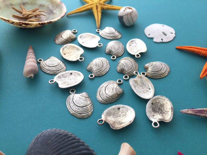 May include: A collection of silver-toned clam shell charms with a textured surface. The charms are arranged on a blue background with seashells and starfish.
