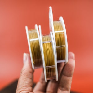 May include: Three spools of gold colored elastic cord, wound on clear plastic spools with white labels. The spools are held in a hand.