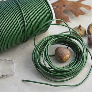 May include: A spool of green leather cord, wound tightly, with a loose end of the cord extending to the right. The cord is lying on a white surface with several acorns nearby.