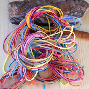 May include: A tangled assortment of colorful cords in shades of red, orange, yellow, green, blue, and purple. The cords are arranged on a light wooden surface, with a rock in the background. The cords are likely for crafting or jewelry making.