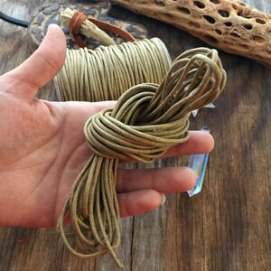 May include: A bundle of olive green cord, knotted and held in a hand, next to a spool of the same cord. The cord appears to be made of a waxed material. The background includes a wooden surface and decorative elements.