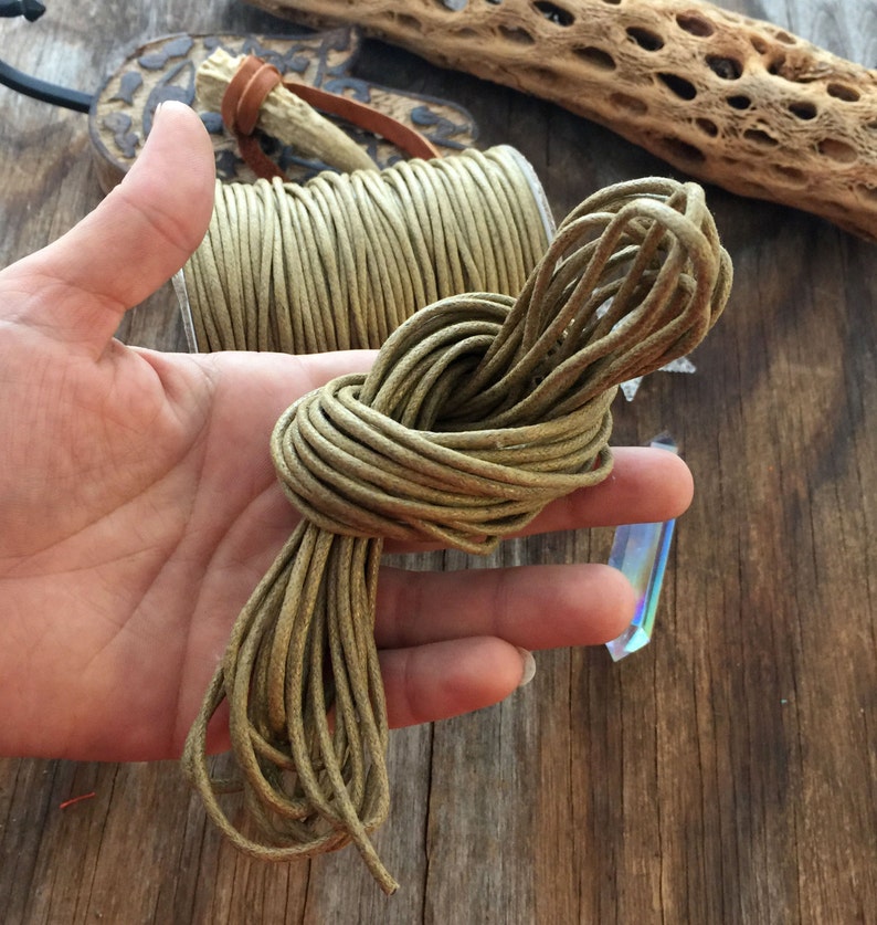 May include: A bundle of olive green cord, knotted and held in a hand, next to a spool of the same cord. The cord appears to be made of a waxed material. The background includes a wooden surface and decorative elements.