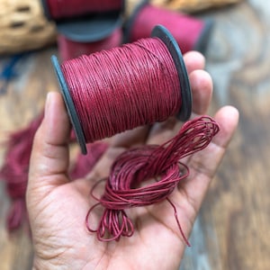 Puede incluir: Primer plano de carretes de hilo de color burdeos. Un carrete se sostiene en una mano, con un pequeño manojo de hilo suelto. Se ven carretes adicionales en el fondo, lo que sugiere un suministro de artesanía o costura.