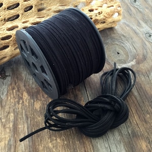 May include: A spool of black suede cord, wound around a black plastic spool. The cord is also shown unwound, lying on a wooden surface.