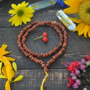 May include: A brown wooden mala bead necklace with a yellow tassel. The necklace is laid out on a dark wood surface with various flowers and crystals around it.