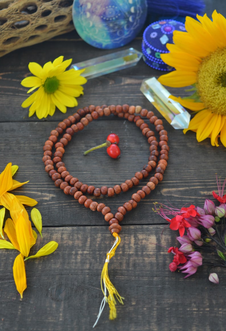 May include: A brown wooden mala bead necklace with a yellow tassel. The necklace is laid out on a dark wood surface with various flowers and crystals around it.