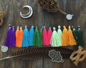 May include: A set of 12 colorful tassels with silver tops. The tassels are arranged in a row on a wooden surface. The tassels are in the following colors: purple, orange, blue, green, pink, light green, orange, and green.
