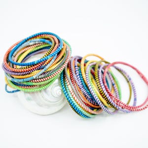 May include: A stack of colorful bracelets made of woven fabric. The bracelets are arranged in a circle and are sitting on a white surface.