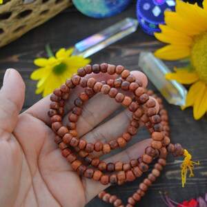 May include: A brown wooden mala bead necklace with a yellow tassel. The necklace is made of small, round beads and is strung on a thin cord. The necklace is lying on a wooden surface.