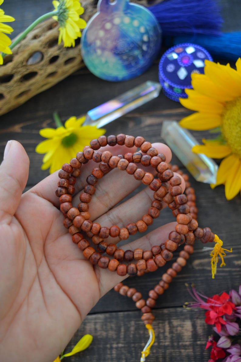 May include: A brown wooden mala bead necklace with a yellow tassel. The necklace is made of small, round beads and is strung on a thin cord. The necklace is lying on a wooden surface.